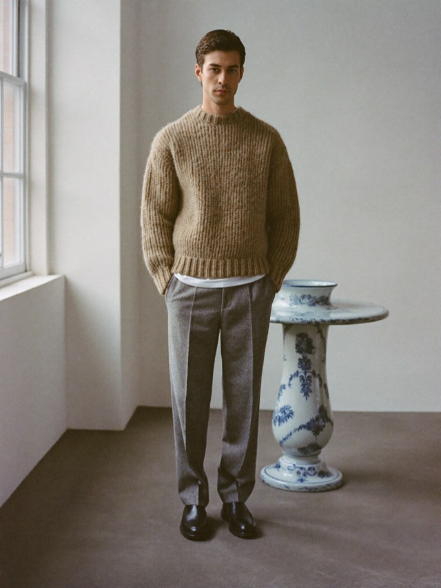 Man in an oatmeal knit sweater and gray wool trousers against a porcelain studio wall